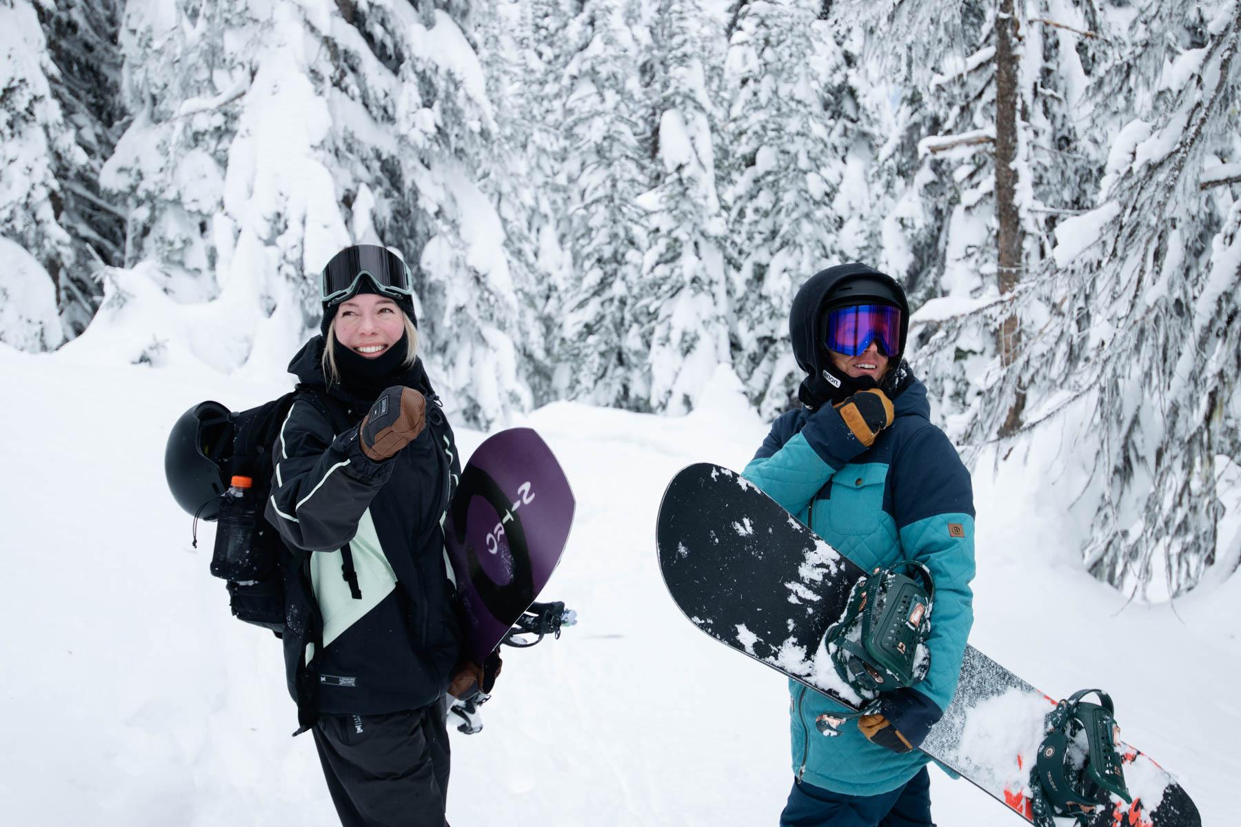 Two snowboarders standing in the trees holding their boards  and smiling at the camera