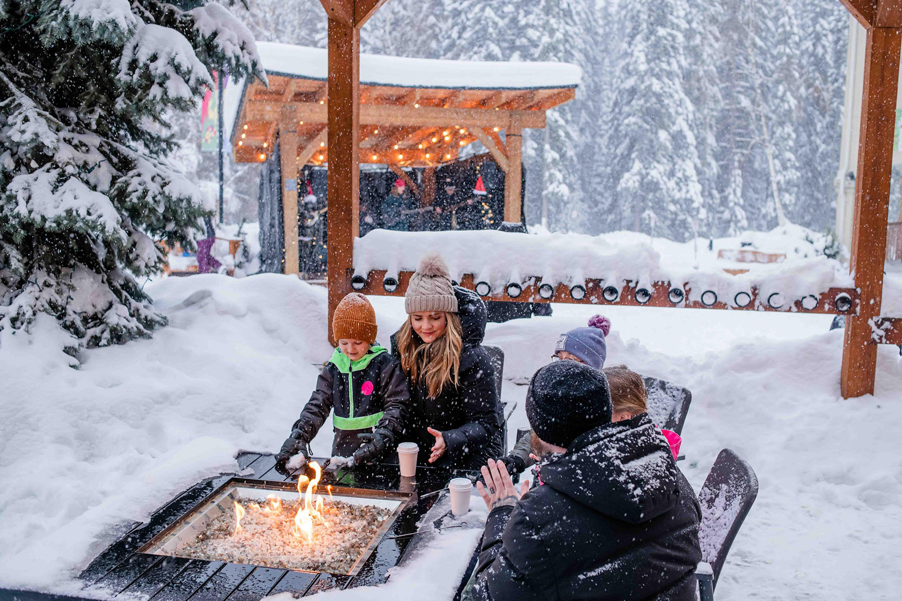 A family sitting around a firepit on a patio with snowfall around them