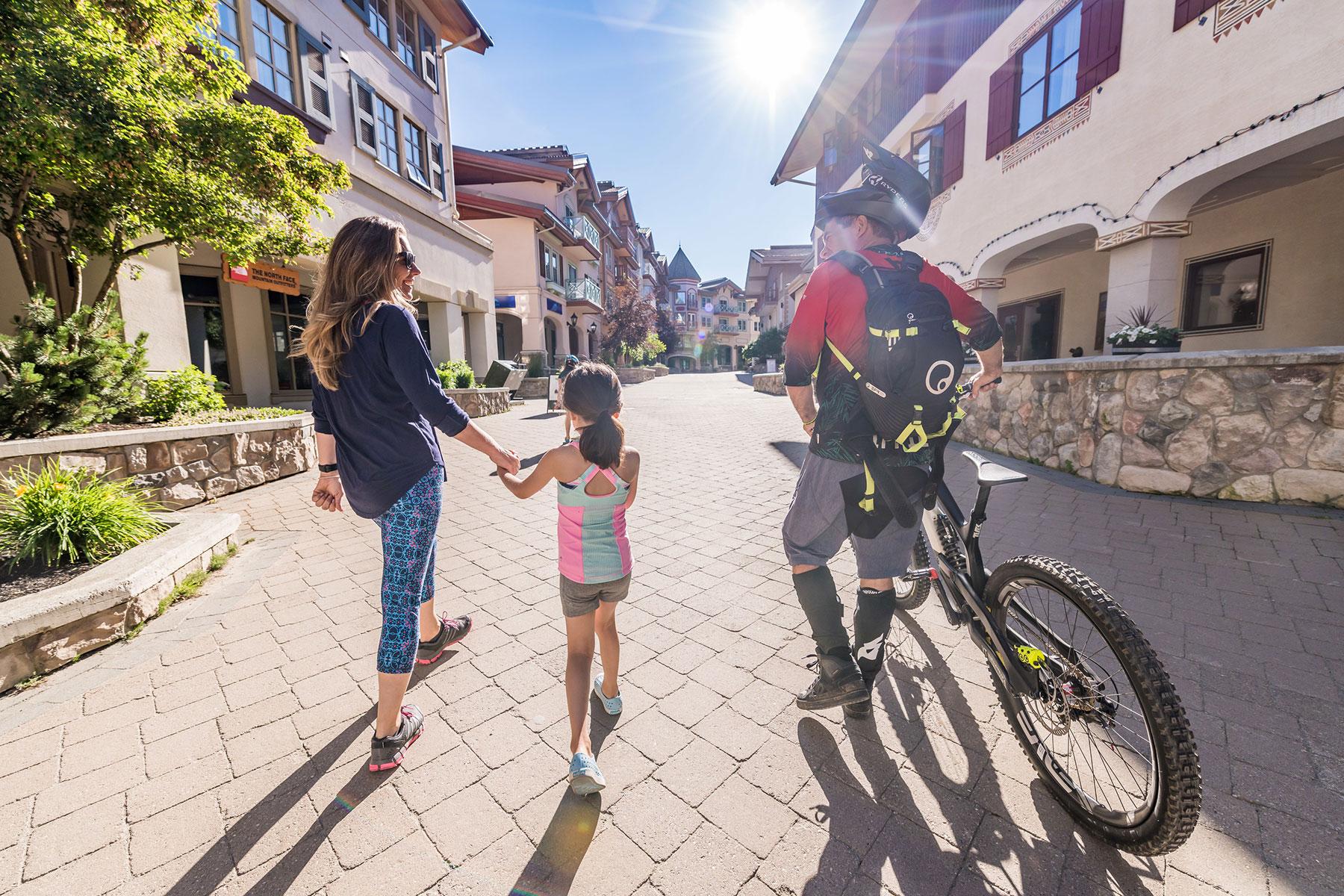 Family walking through Sun Peaks village on a bright summer day, father with a mountain bike.