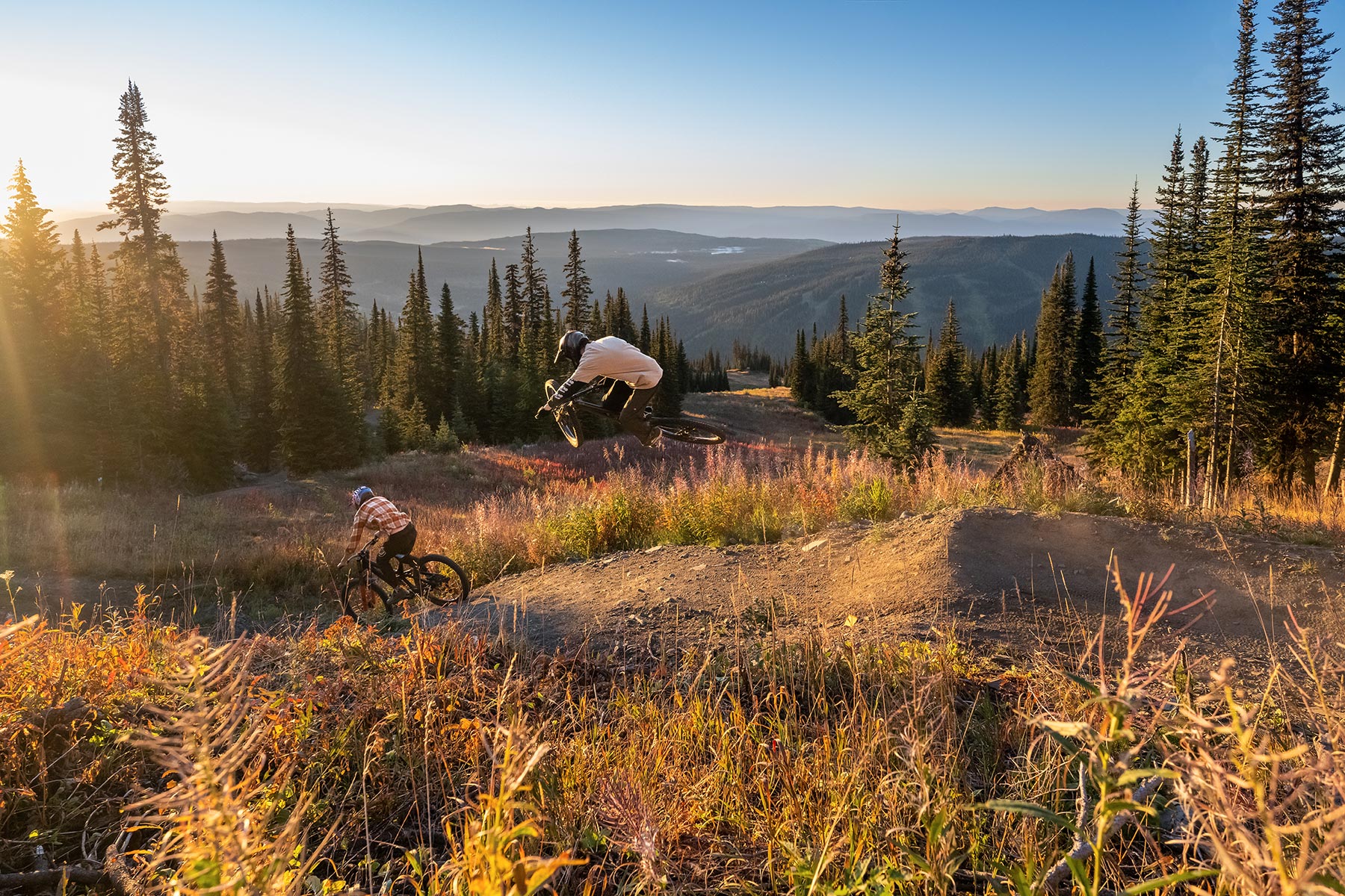 Two bikers on a trail and one if hitting a jump with the sunsetting in the background