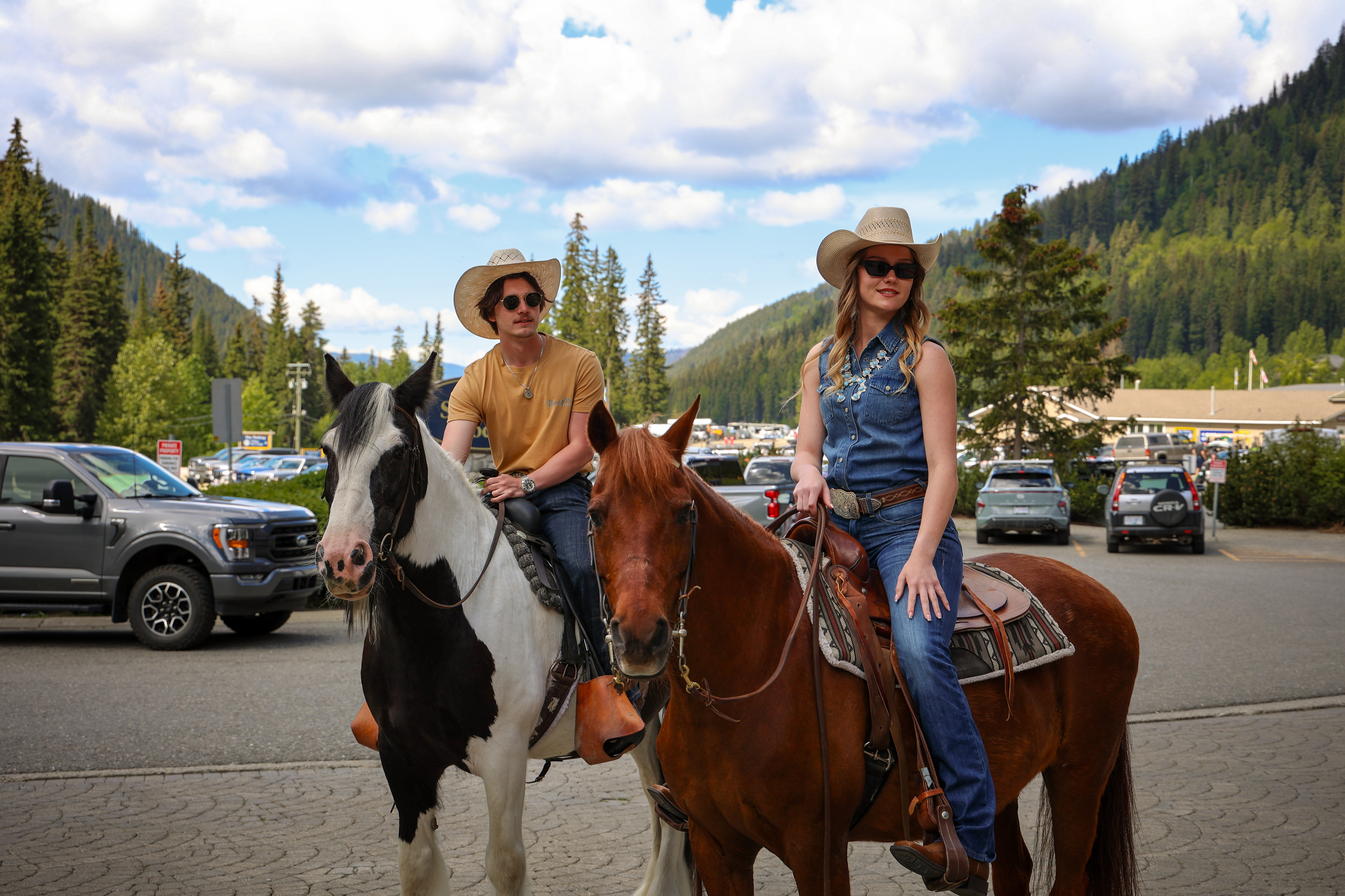 A man and a woman in country clothing and cowboy hats, riding horses in Sun Peaks village.