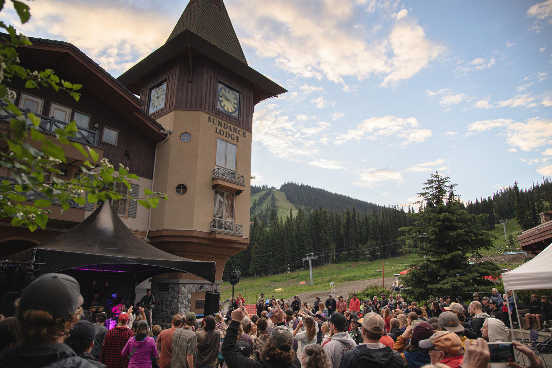 Crowd watching a band at the Clock Tower Stage at Sun Peaks on Canada day with mountains and a cloudy blue sky visible behind.