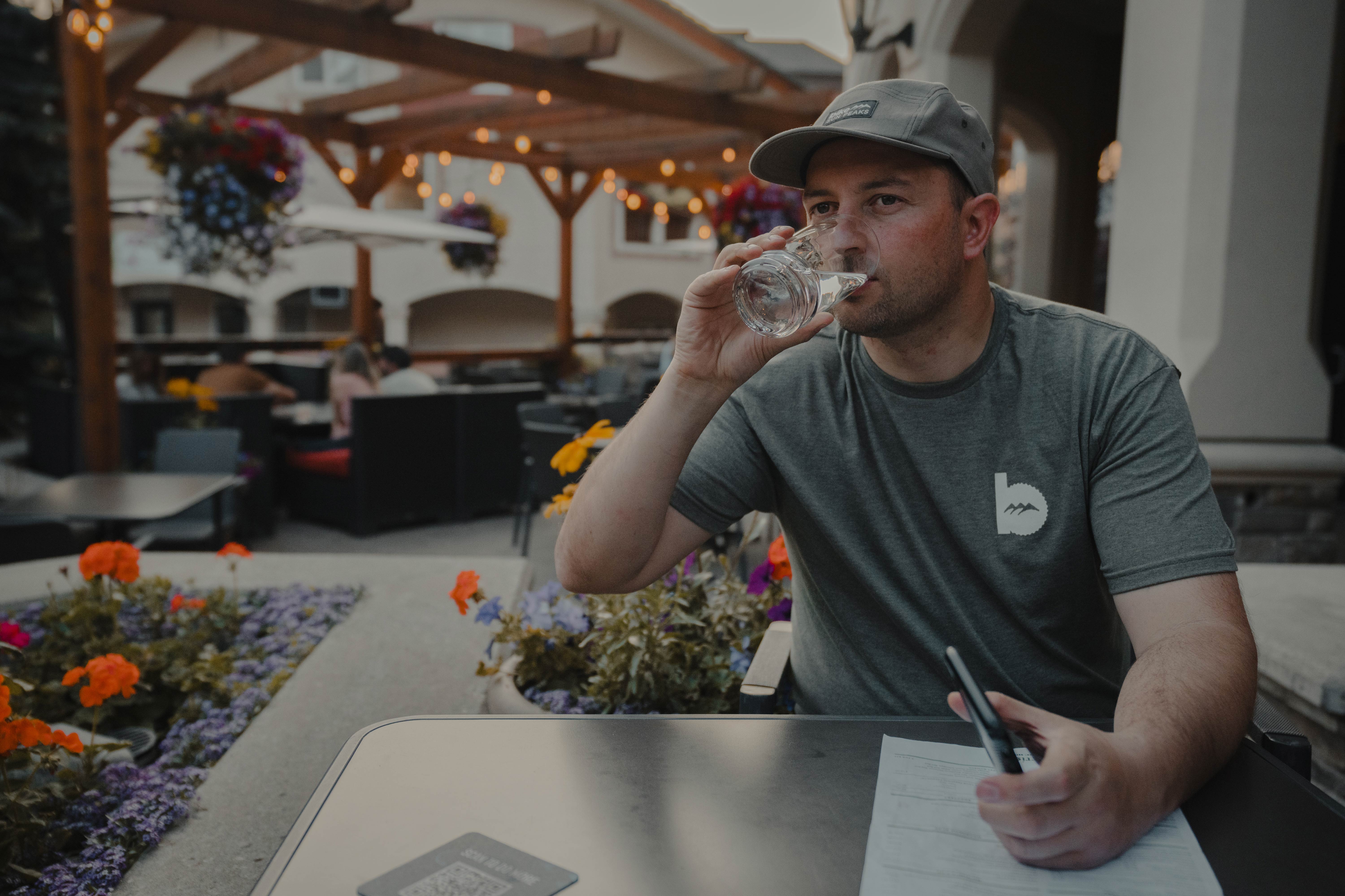 Man in a grey shirt drinking a glass of water under string lights on a summer patio. 