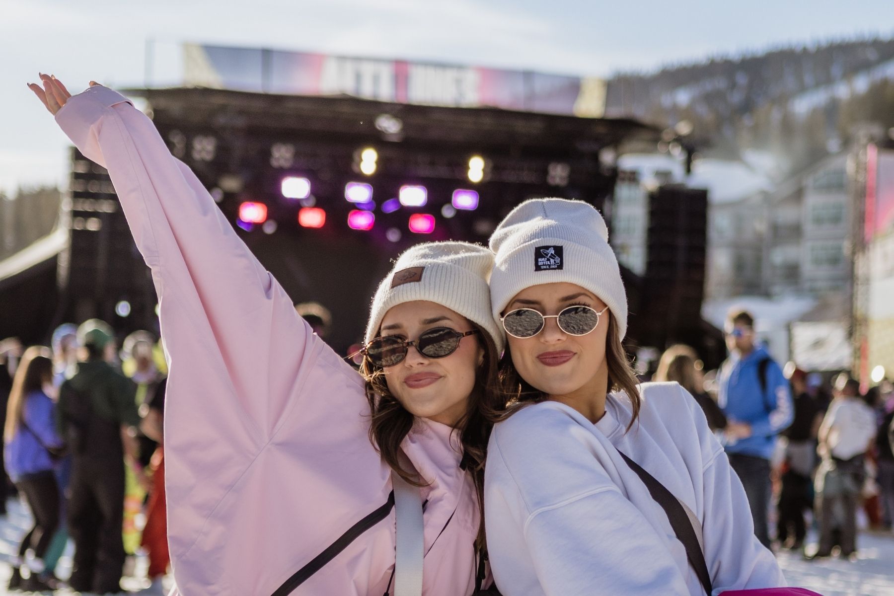 Two girls in pink and white sweaters and hats with sunglasses in front of AltiTunes stage