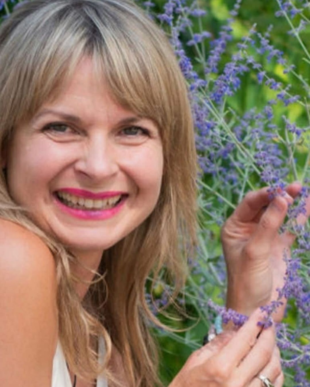 Angie Szerepi in a green field holding purple flower, smiling with bright pink lipstick.