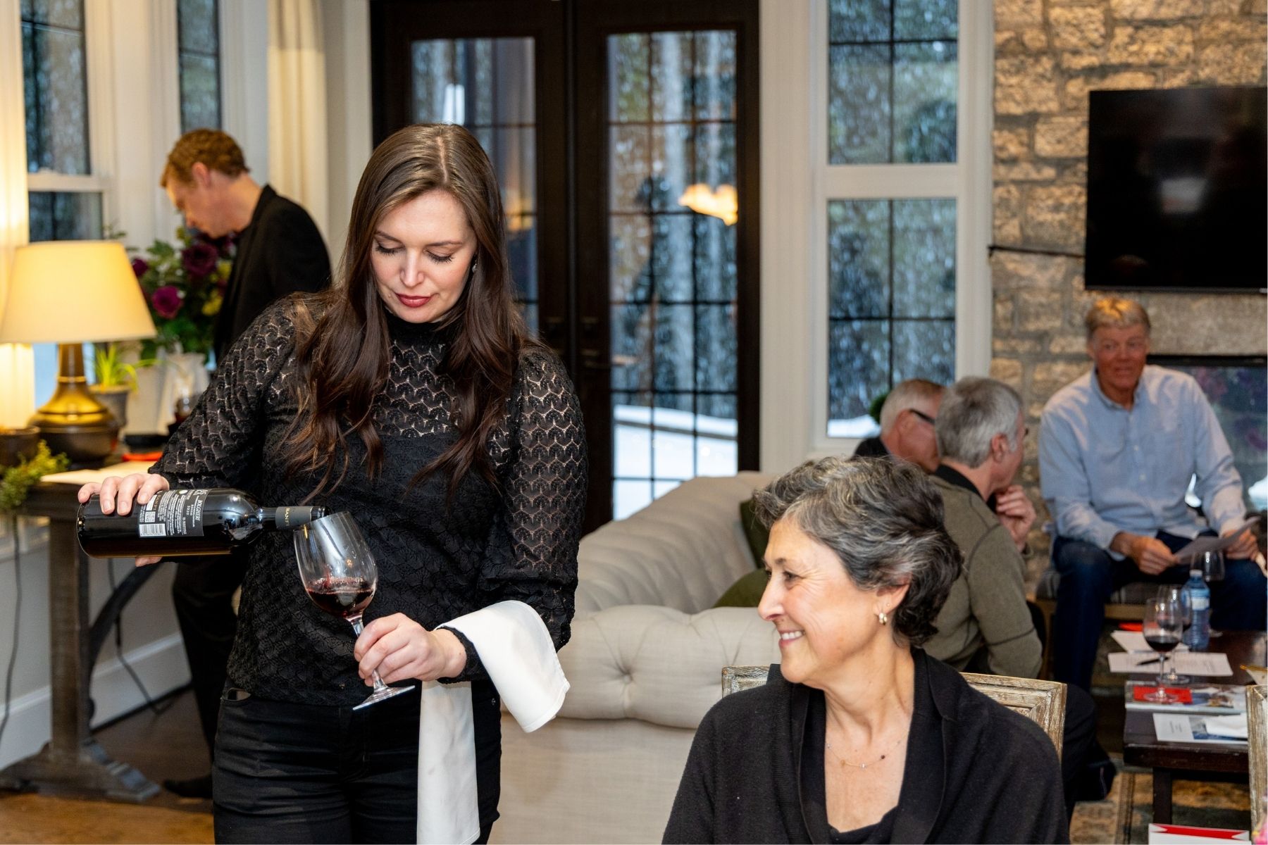 Woman in a black dress pouring wine for a smiling woman at an elegant chalet party.
