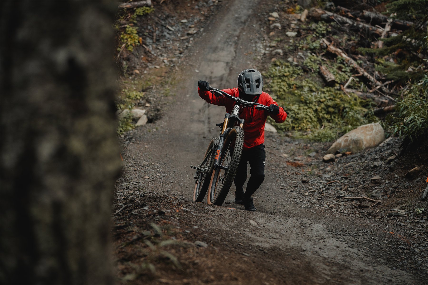 Mountain biker in full face helmet and red jacket pushing his bike up a hill