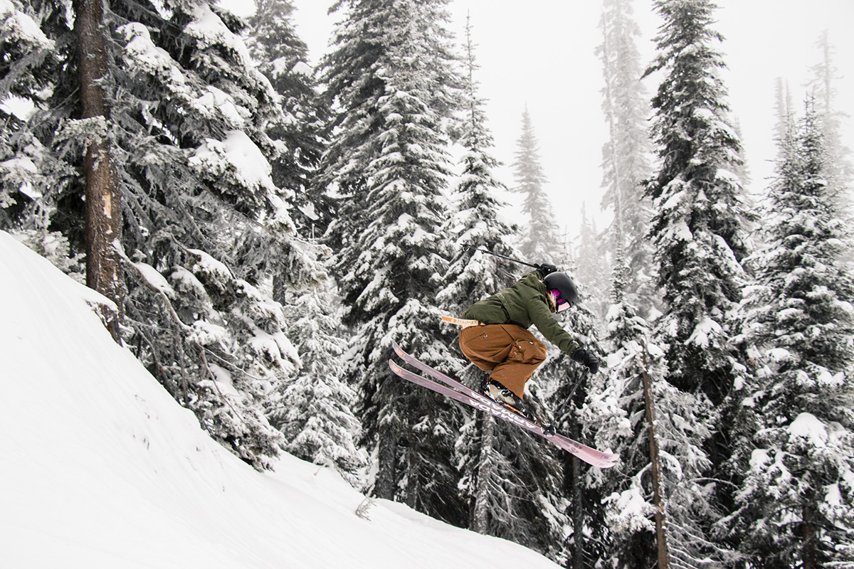 Skier, Gabi Dinn, drops off a snowy side hit in front of trees.