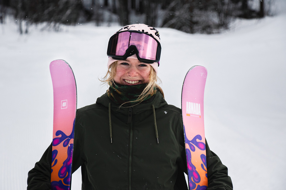 Gabi Dinn smiles for the camera wearing a pink hat with pink skis in each hand.