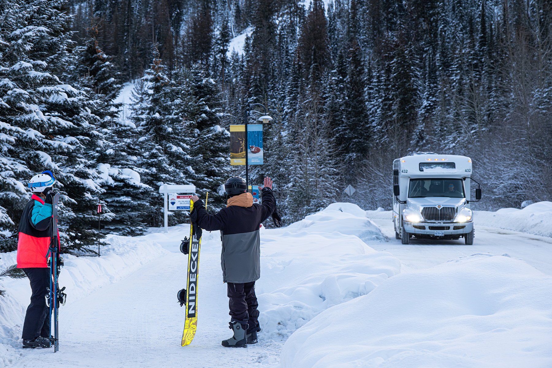A skier and snowboarder waiving at the incoming in-resort shuttle, on a snowy street with trees and snowbanks