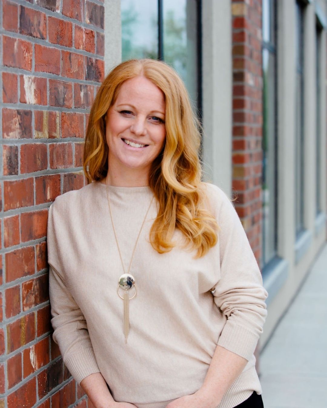 Joanne Parker, with curling red hair and a light tan shirt, leaning against a brick wall and smiling.