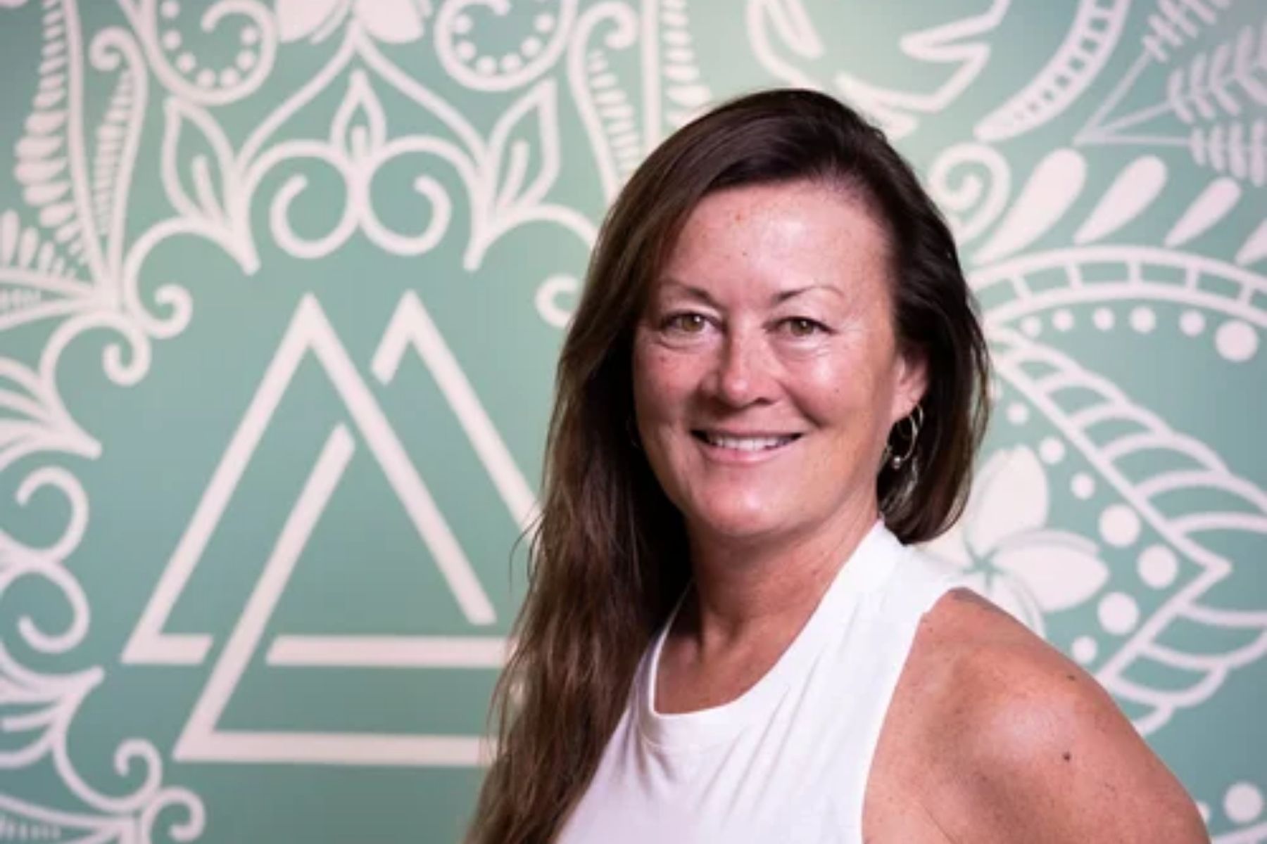 Kelly Craig, yoga teacher smiling in a white tank top in front of a blue and white decorated wall.