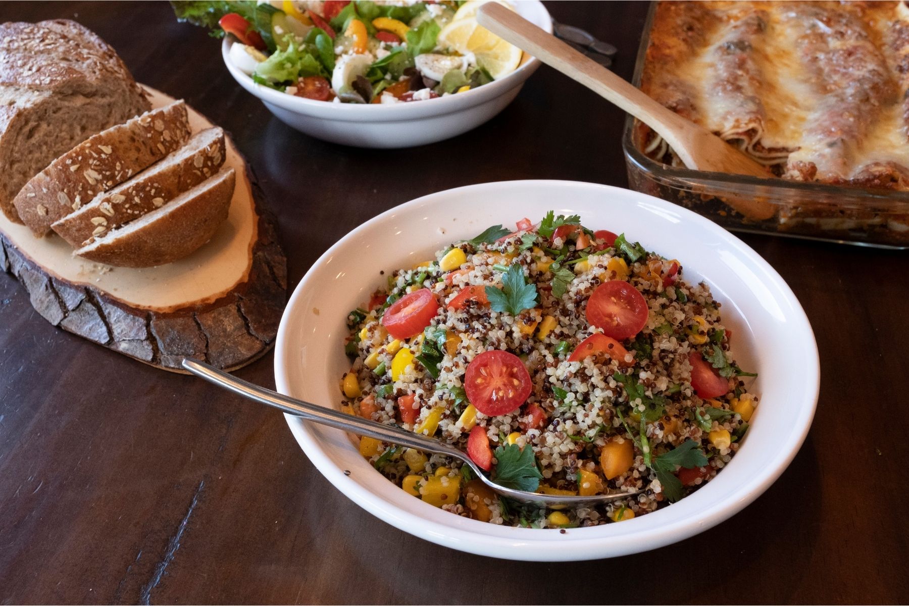 A bowl of healthy quinoa salad, a lasagna, a fresh summer salad, and a loaf of bread on a wooden table.
