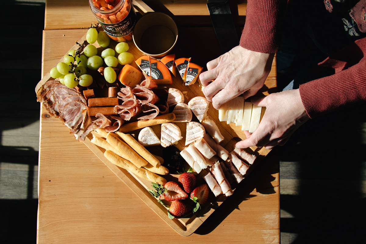 Hands preparing a charcuterie board from Ohana Deli Market in Sun Peaks, with cheese, meat, bread, and fruit.