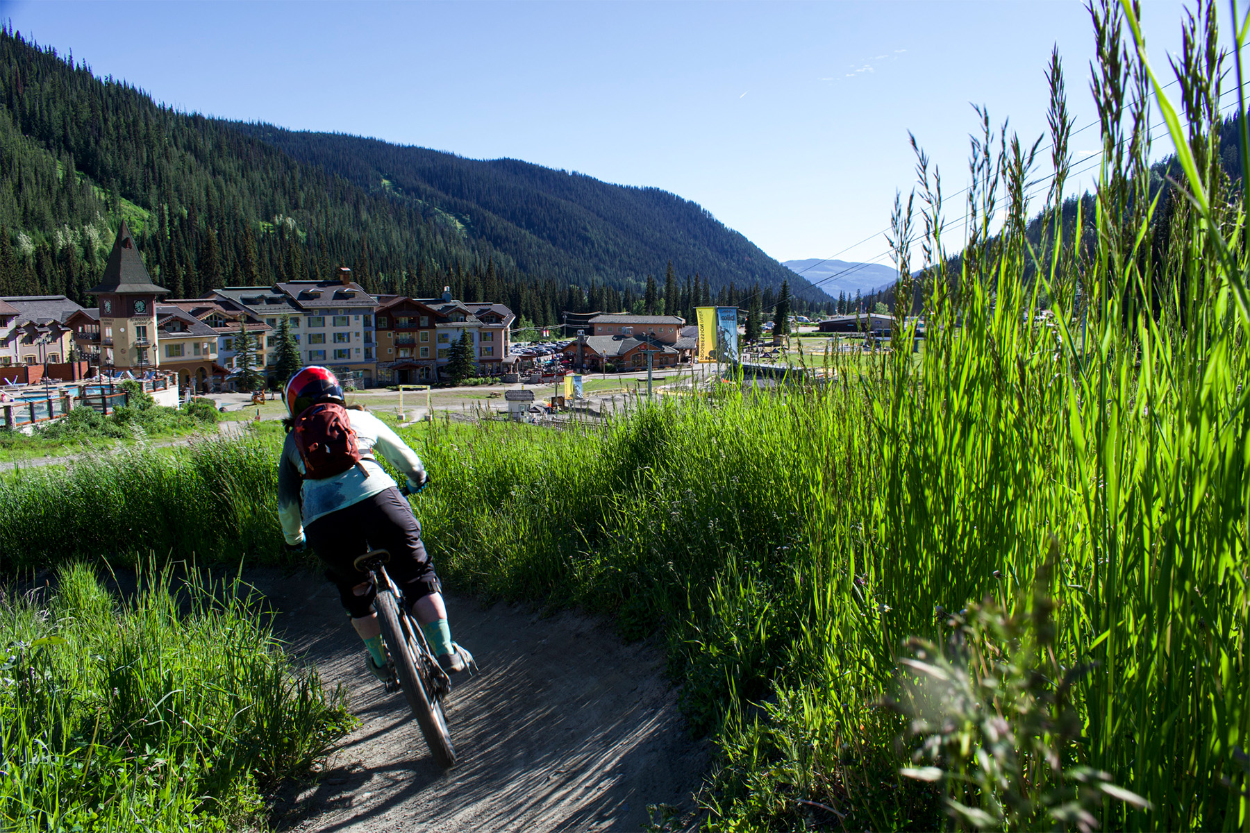 Biker in Progression Park, on a trail between tall grass in front of an alpine village.