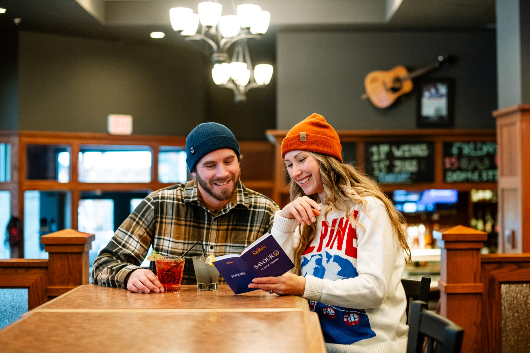 Couple in beanies smiling in a cozy restaurant with cocktails and the Savour Passport.