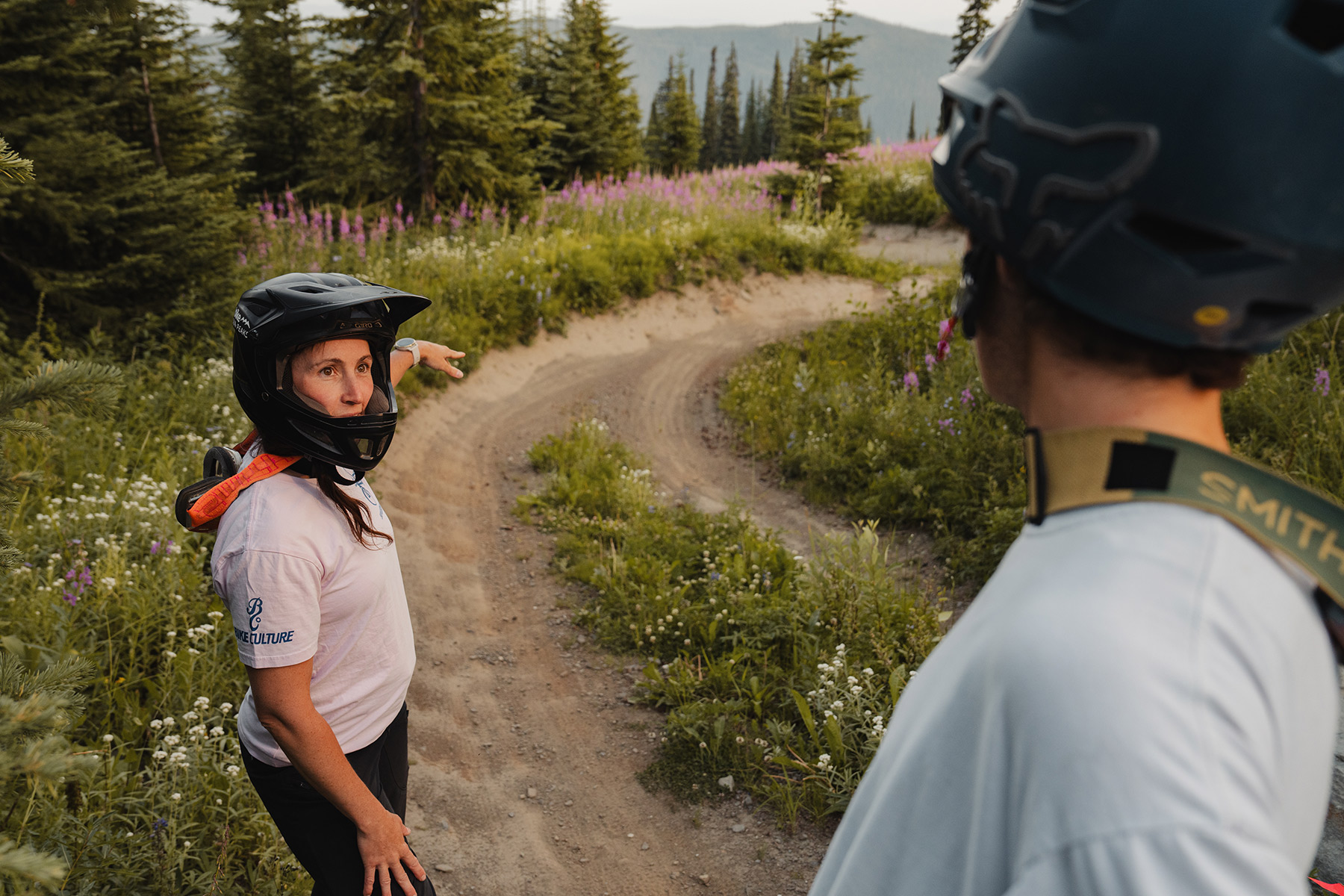 Two mountain bikers stand and look at a berm surrounded by pink wildflowers.