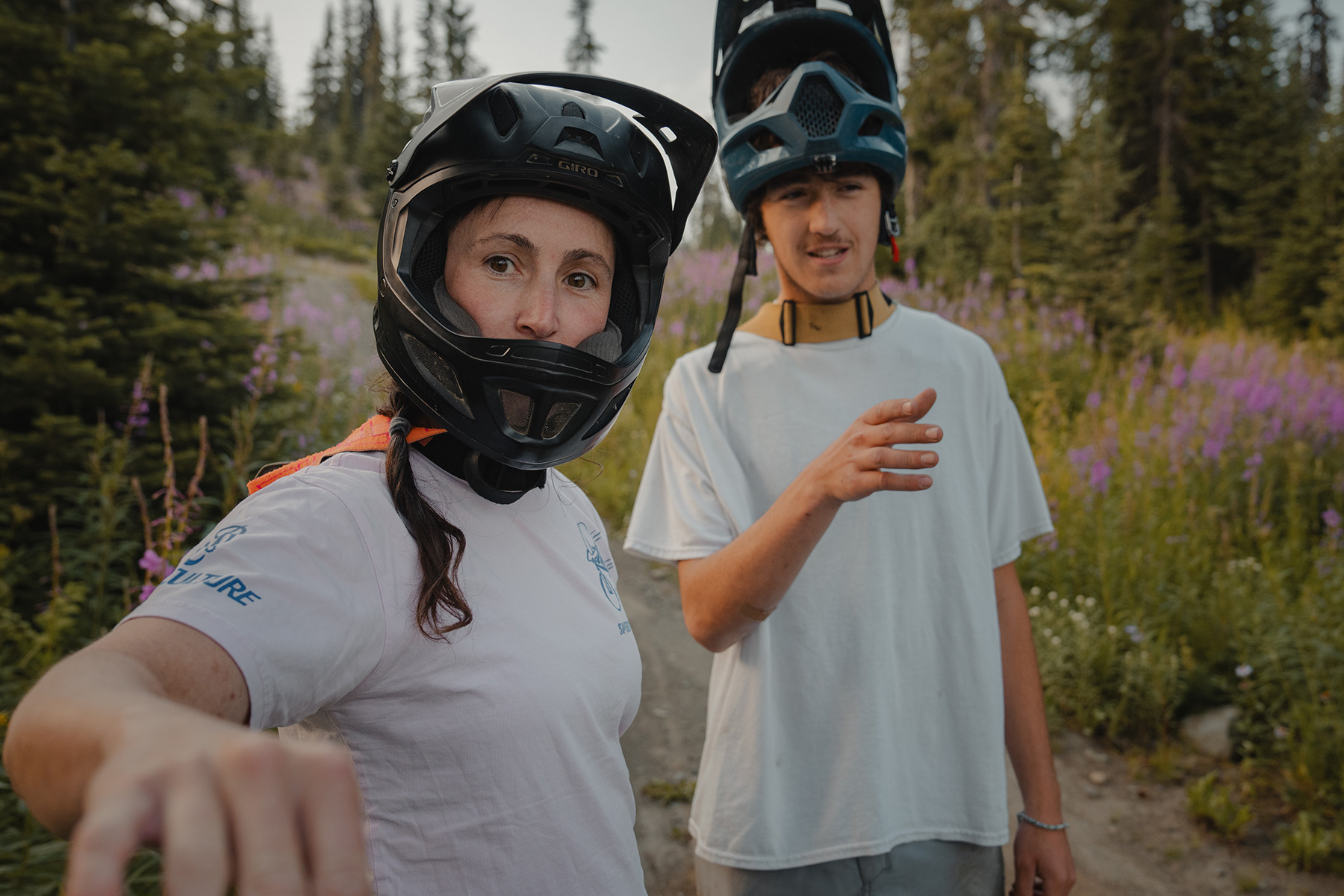 Two mountain bikers in full face helmets look past the camera at a trail.