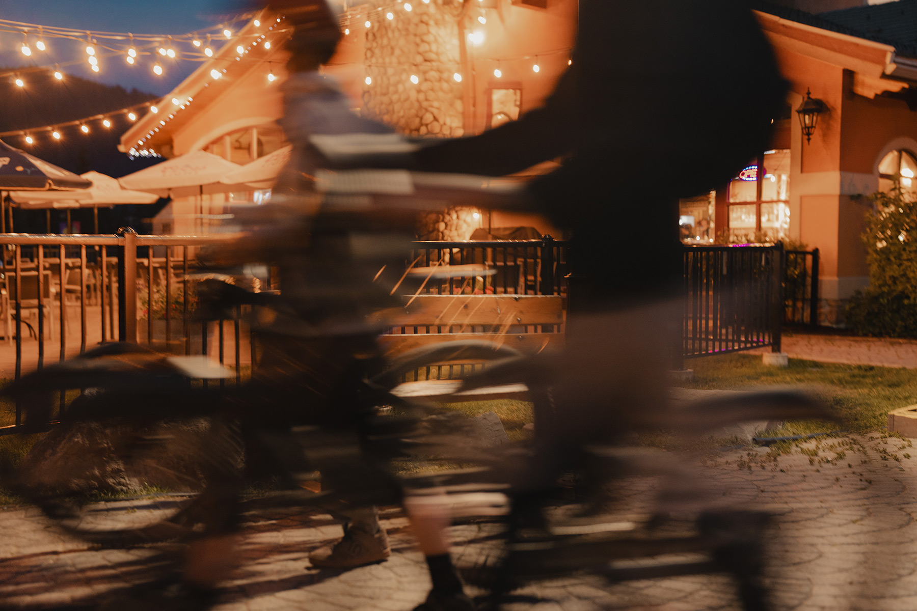 Motion blurs as three bikers walk in front of a glowing outdoor patio at night.