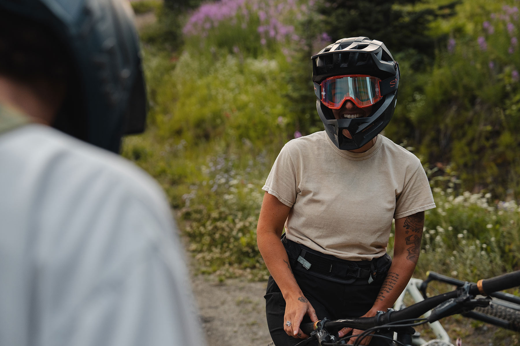 A woman in a full face mountain bike helmet and goggles sits on her bike and laughs at another person.