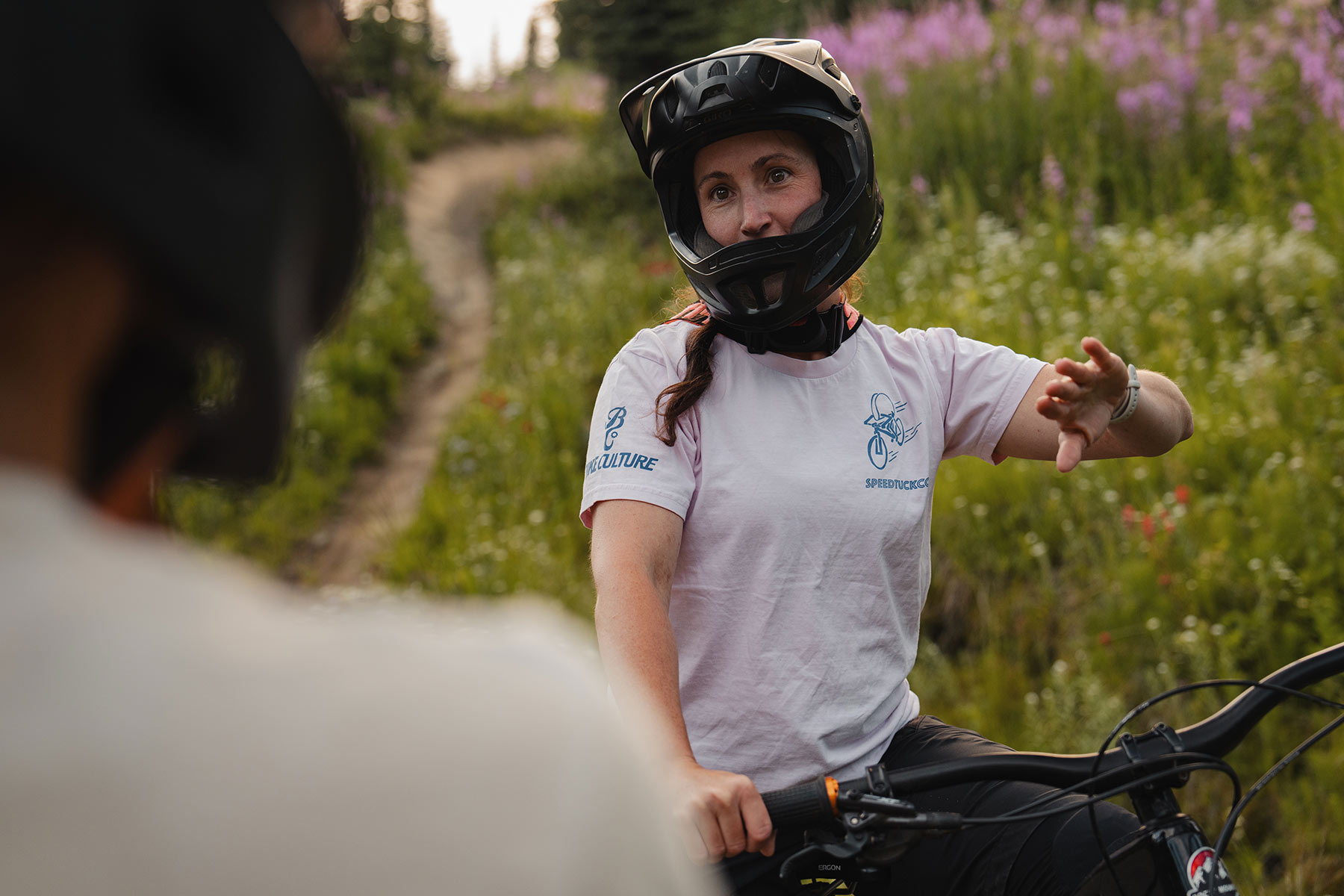 A woman in a full face mountain bike helmet sits on her bike and motions to another person.