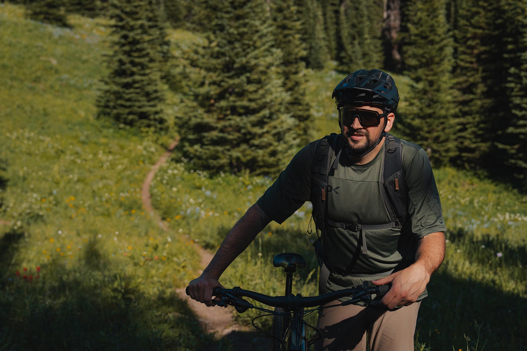 A mountain biker stands beside his bike with a singletrack alpine trail behind him.