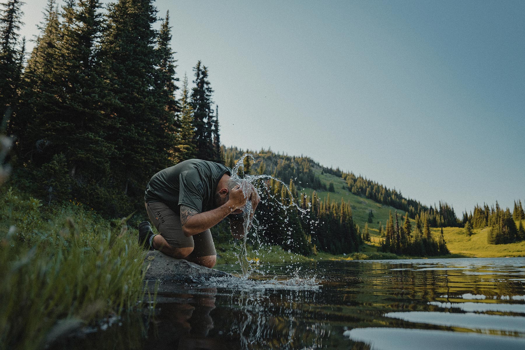 A man kneels by the side of an alpine lake and splashes water over his head.