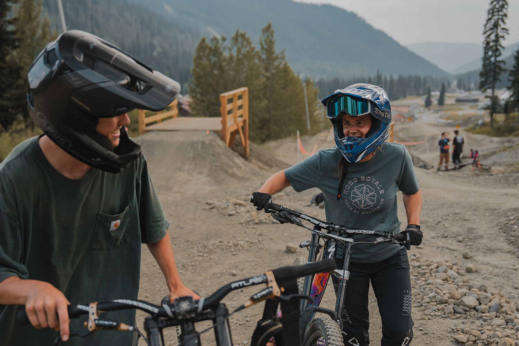 A women in a full-face DH helmet smiles at another mountain biker as they push their bikes up a trail with wooden features.