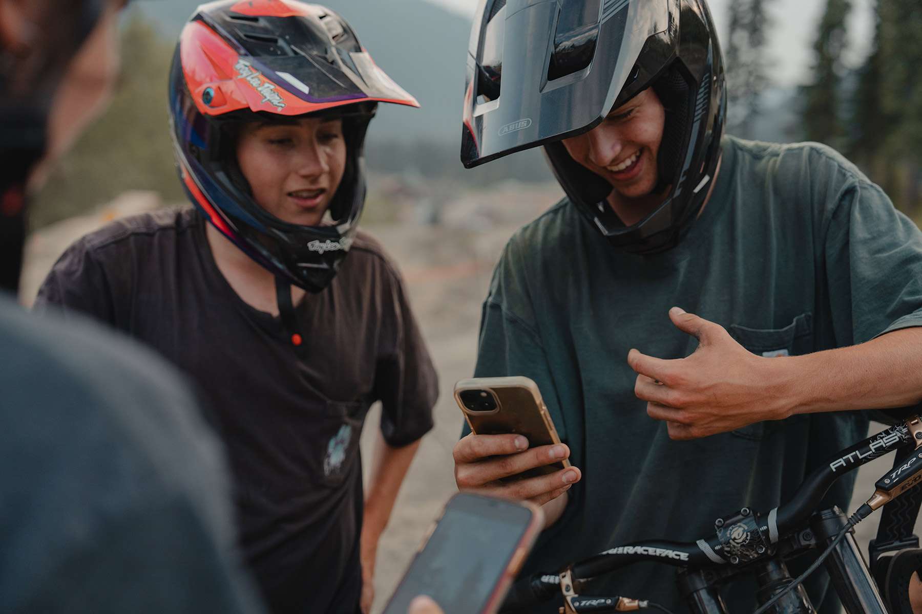 Two mountain bikers in full-face DH helmets look and smile at a phone.
