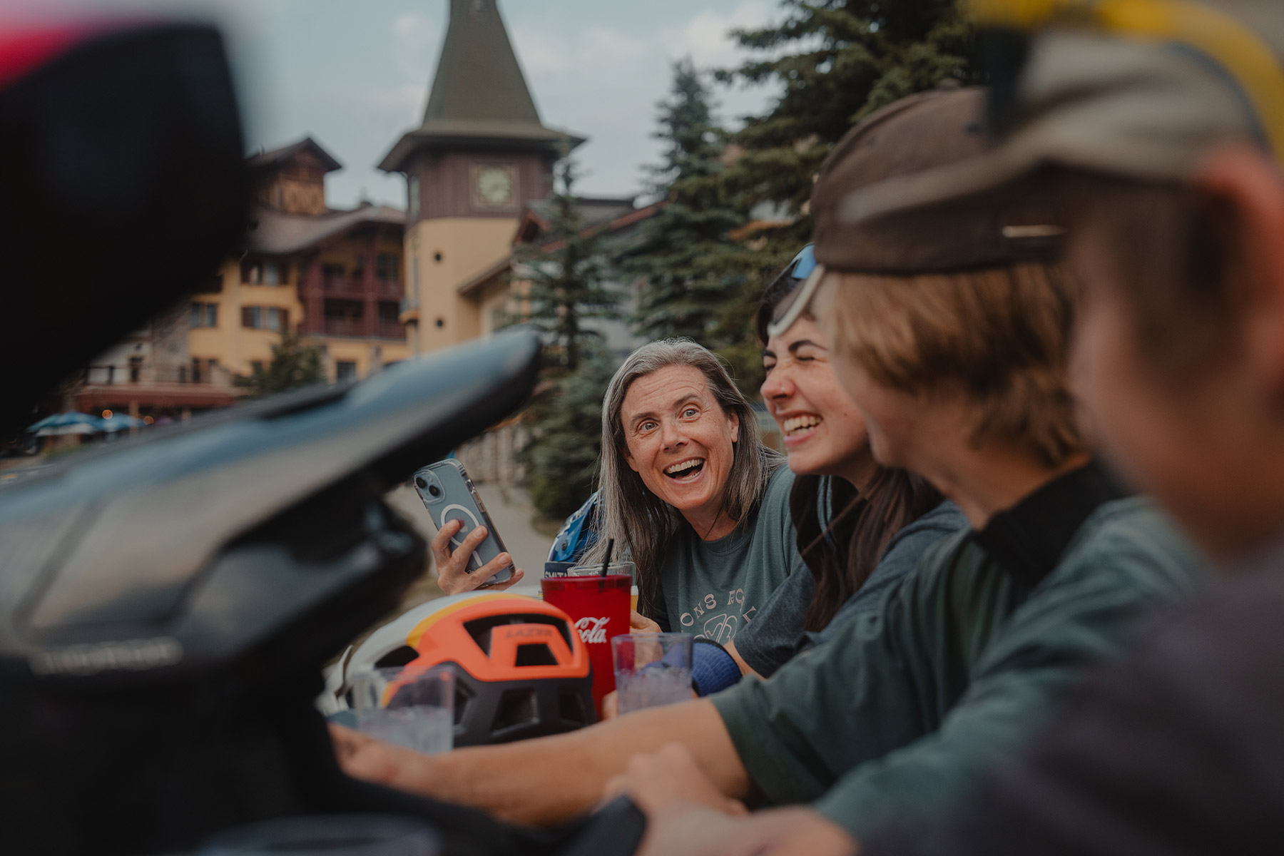 Four people seated at an outdoor patio bar table in front of the Sun Peaks Village clock tower. All smile and laugh at each other.