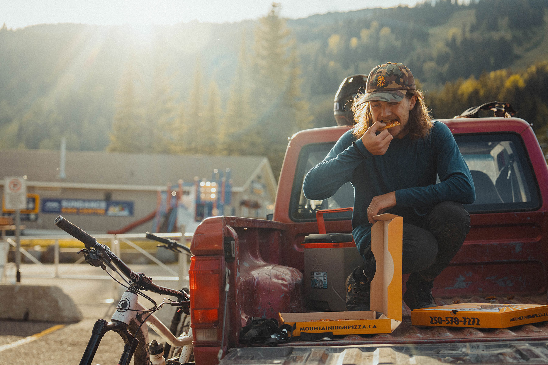 A man in a blue shirt squats in the back of a pickup truck, eating the last piece of pizza from an orange box.
