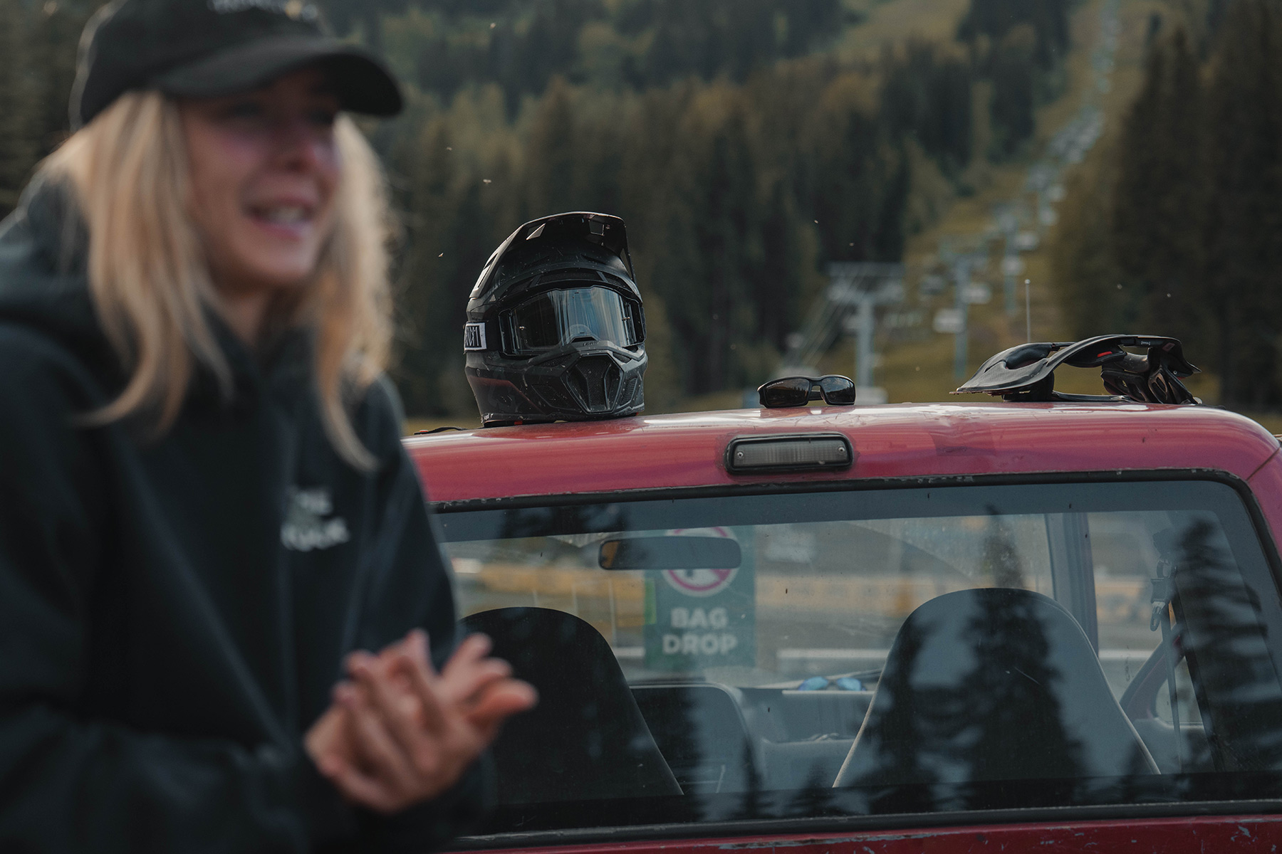 Bike gear sits on top of the red pickup truck with a chairlift in the background and a women in the foreground.