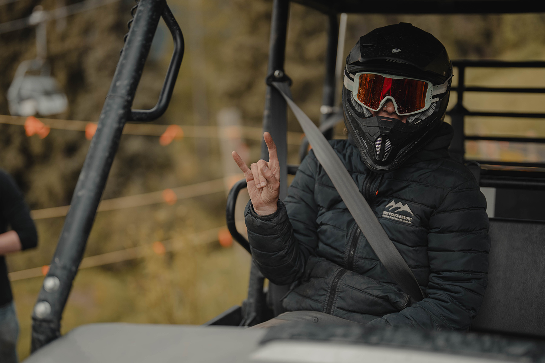 A trail builder wearing a helmet and goggles give throws up a devil horns sign with her hand while riding in a side-by-side atv.