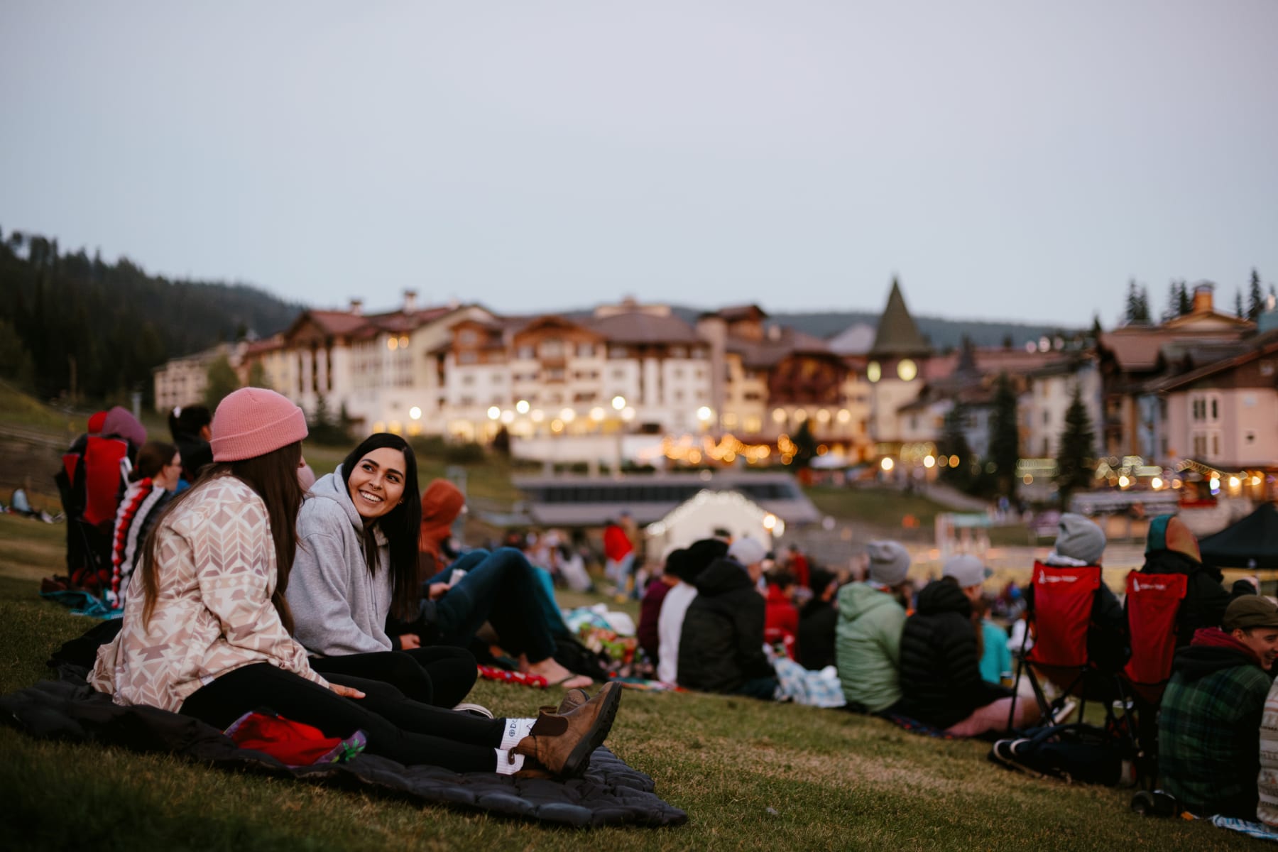 Live music in Sun Peaks at the slopeside stage at twilight. Two girls smiling in foreground on camp blankets at twilight