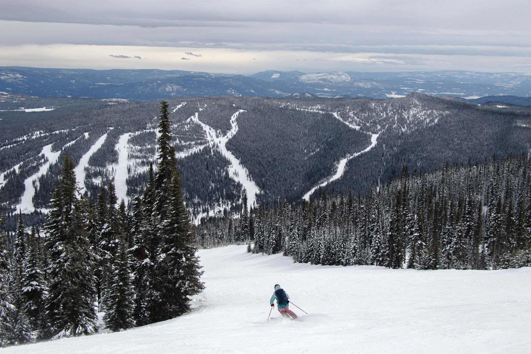 Alpine skier skiing alone, looking out over a ski mountain with the Shuswap Highlands in the background.