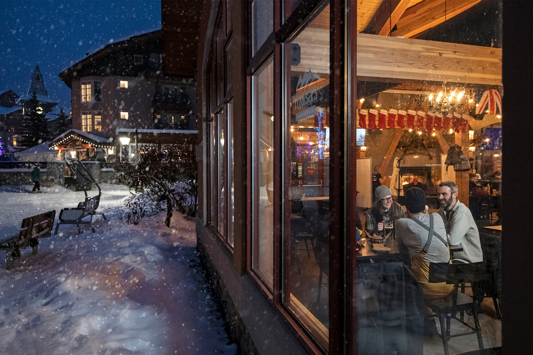 Snow falls at night outside warmly lit Masas Bar in Sun Peaks Village