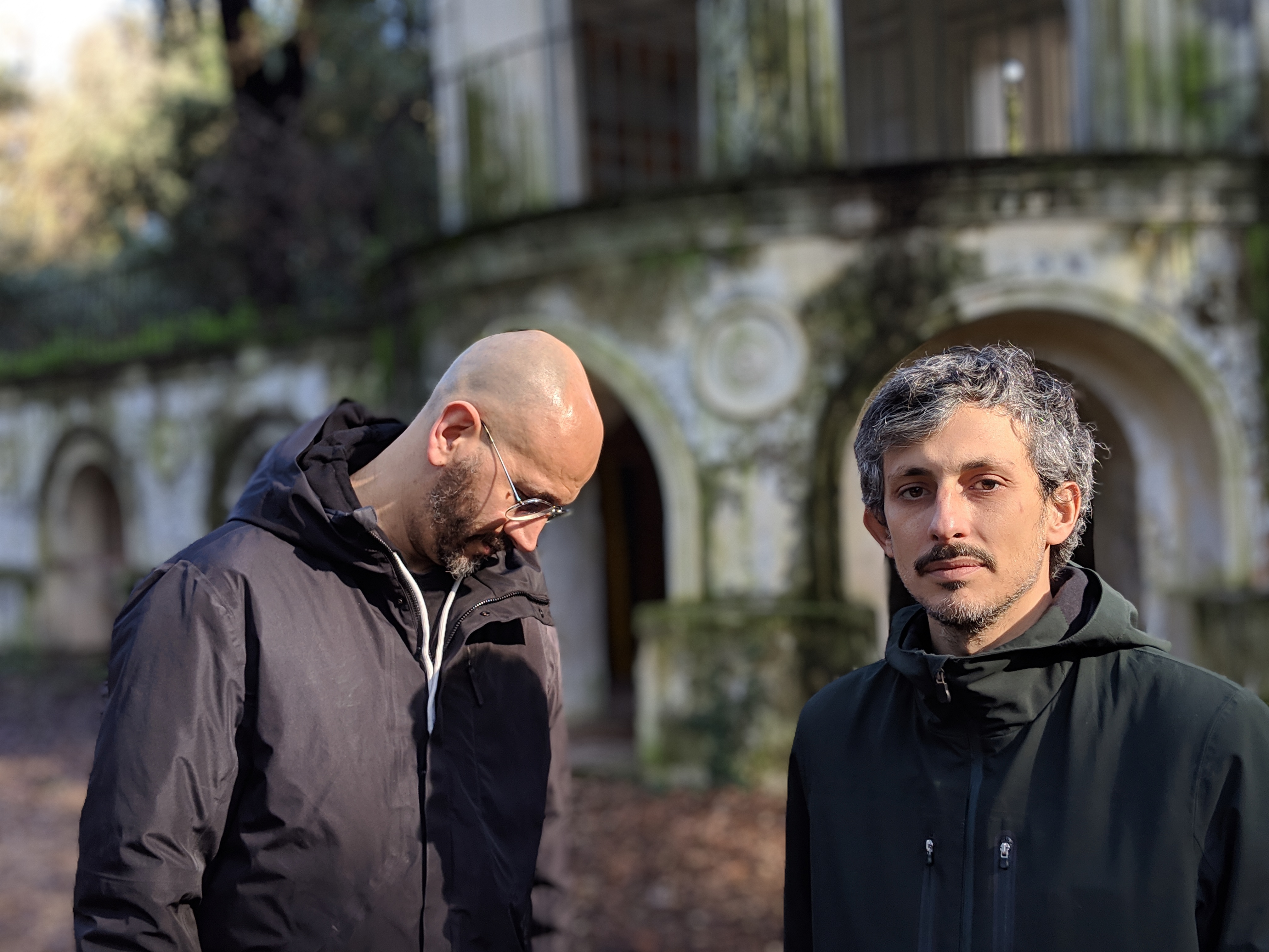 Two men in raincoats, one looking at the camera and one looking down. Old white building behind with ornate arches and green vines around them.
