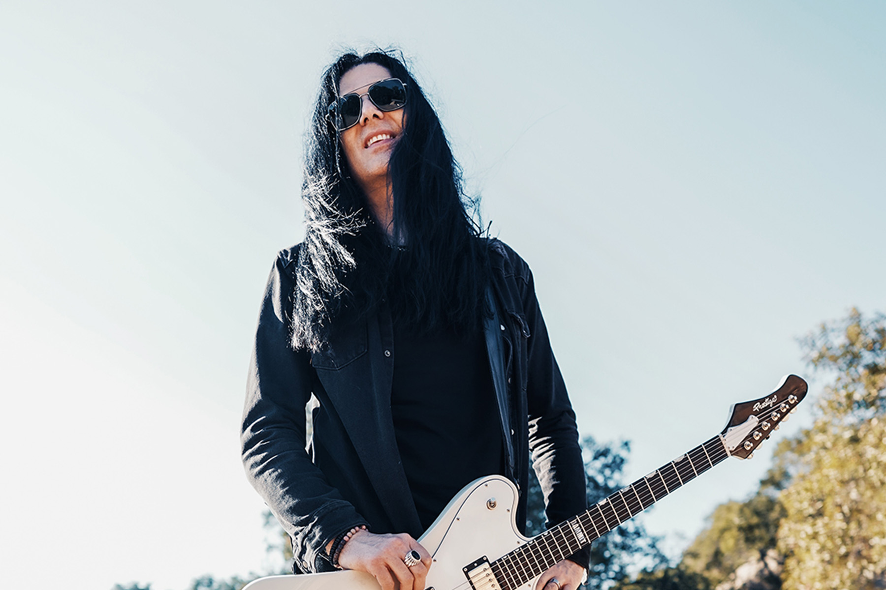 Todd Kerns of the band Toque, dressed in black and holding a guitar against a backdrop of blue sky and trees.