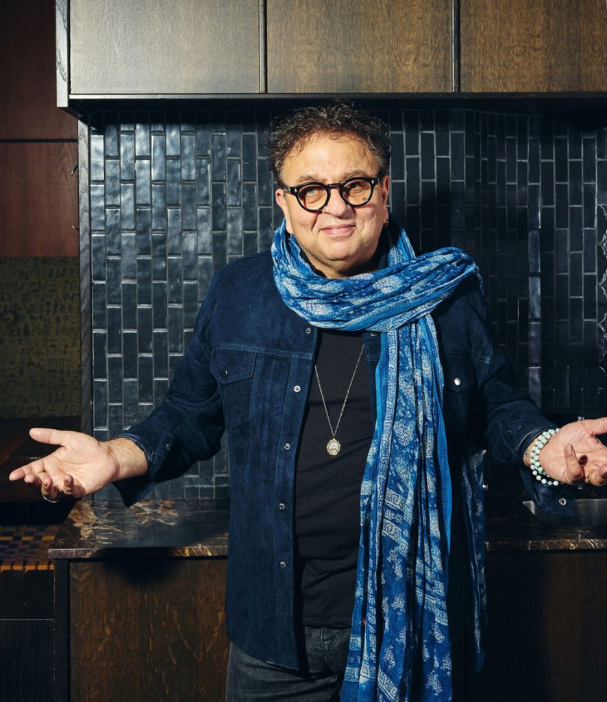 Vikram Vij in a kitchen with a blue scarf and hands up on either side.