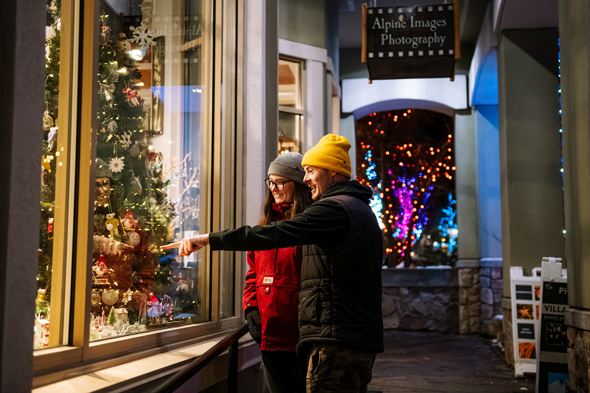 A man points out to a woman a Christmas tree in the window of Alpine images photography in a lit-up Sun Peaks village at night.