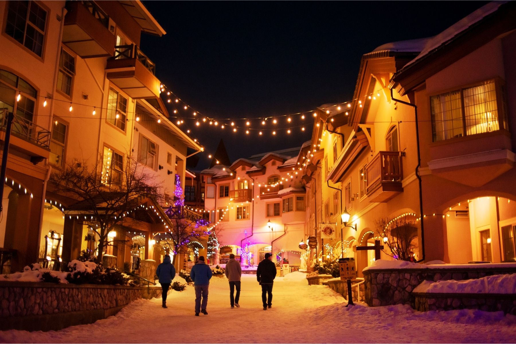 A cozy winter village with sparkling lights hanging overhead and people walking through.