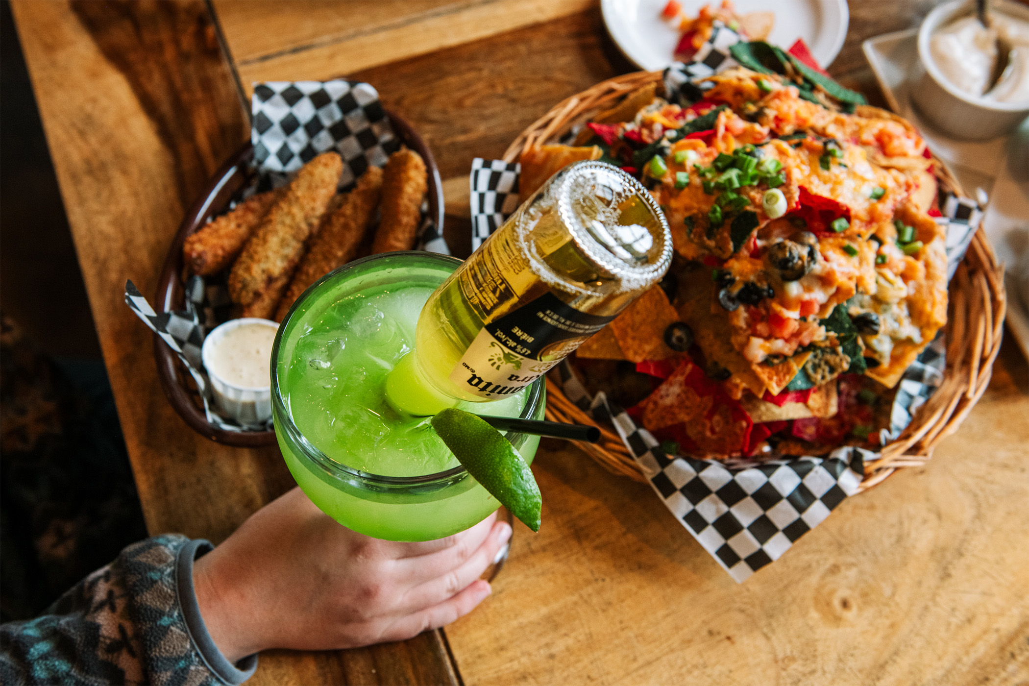 A platter of cheesy nachos, a plate of fried pickles and dip, and a green cocktail with a Corona beer sticking out of the top on a table at après.
