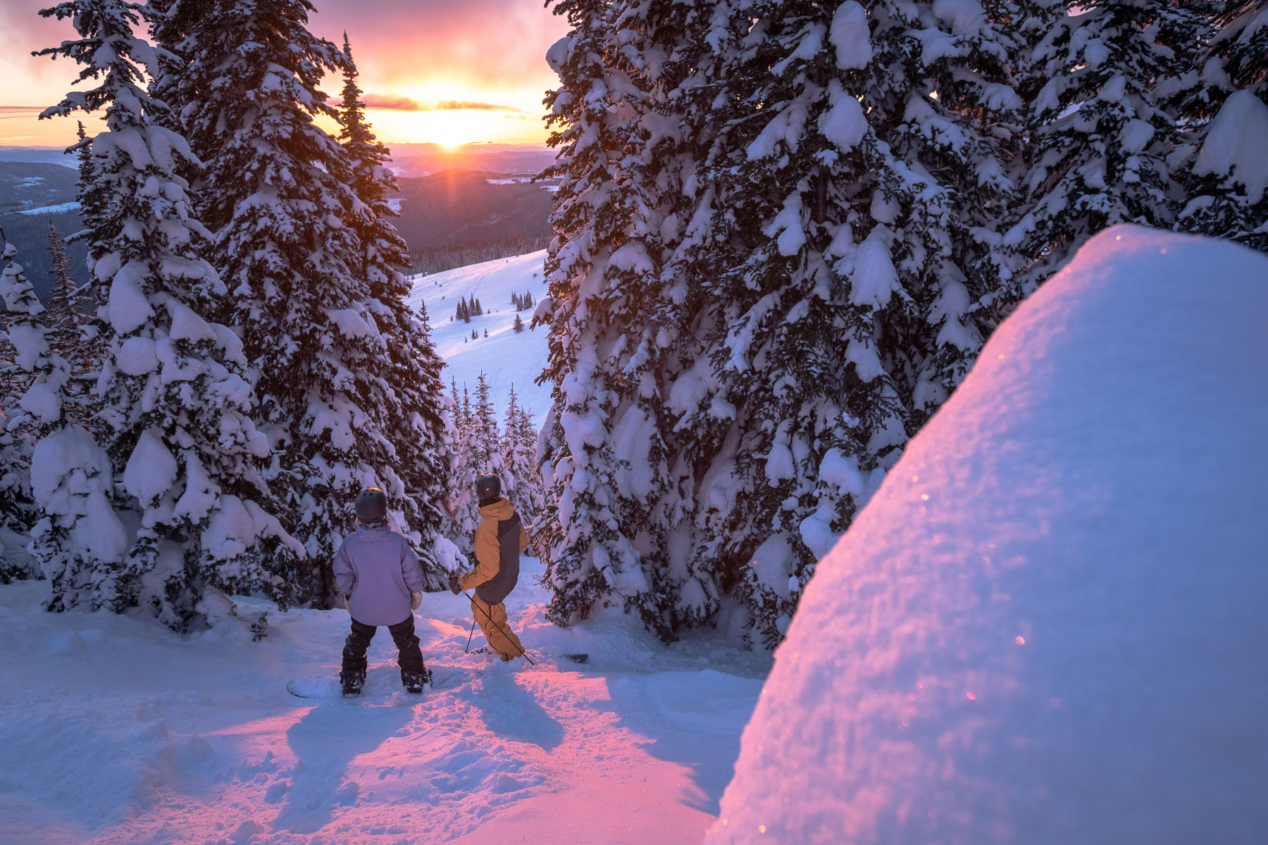 a skier and snowboarder watch a sunset over the sun peaks alpine from a gap in the trees