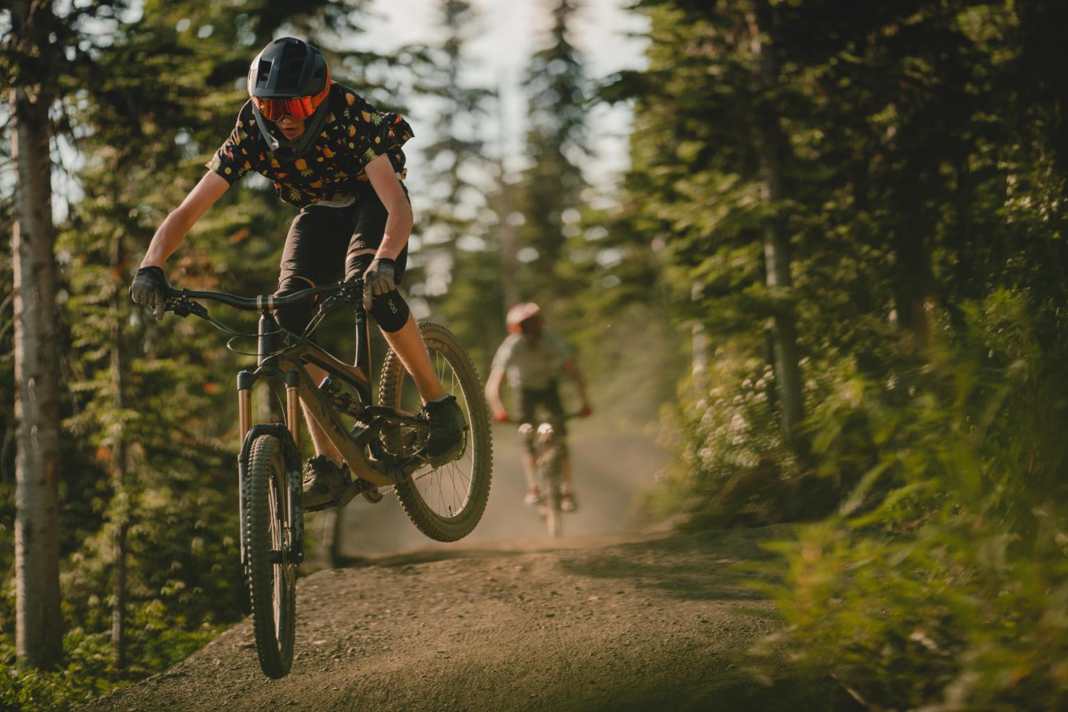 Two bikers hitting a jump with trees surrounding them 