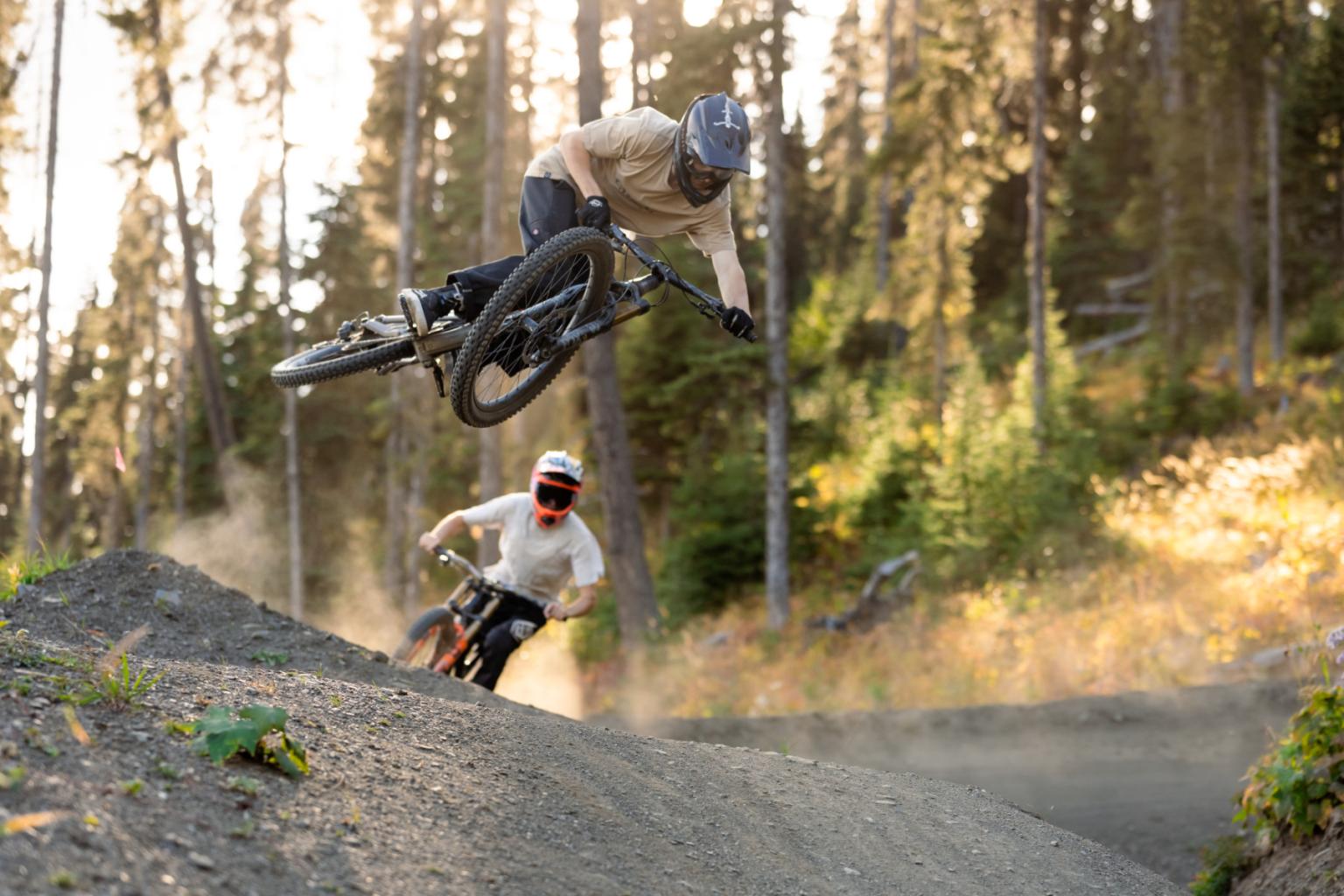 Two bikers hitting a jump with trees surrounding them 
