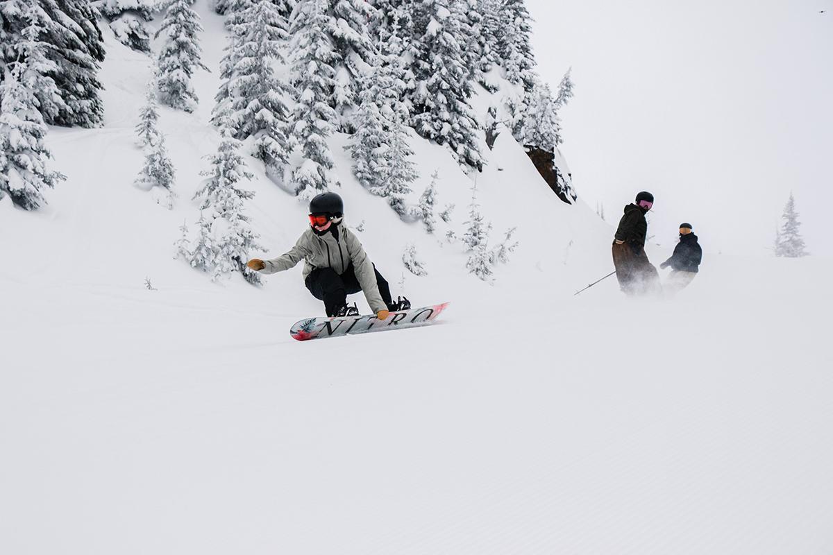 Snowboarder grabs toe-side of board while carving a groomed run, followed by a skier riding switch and a second snowboarder.