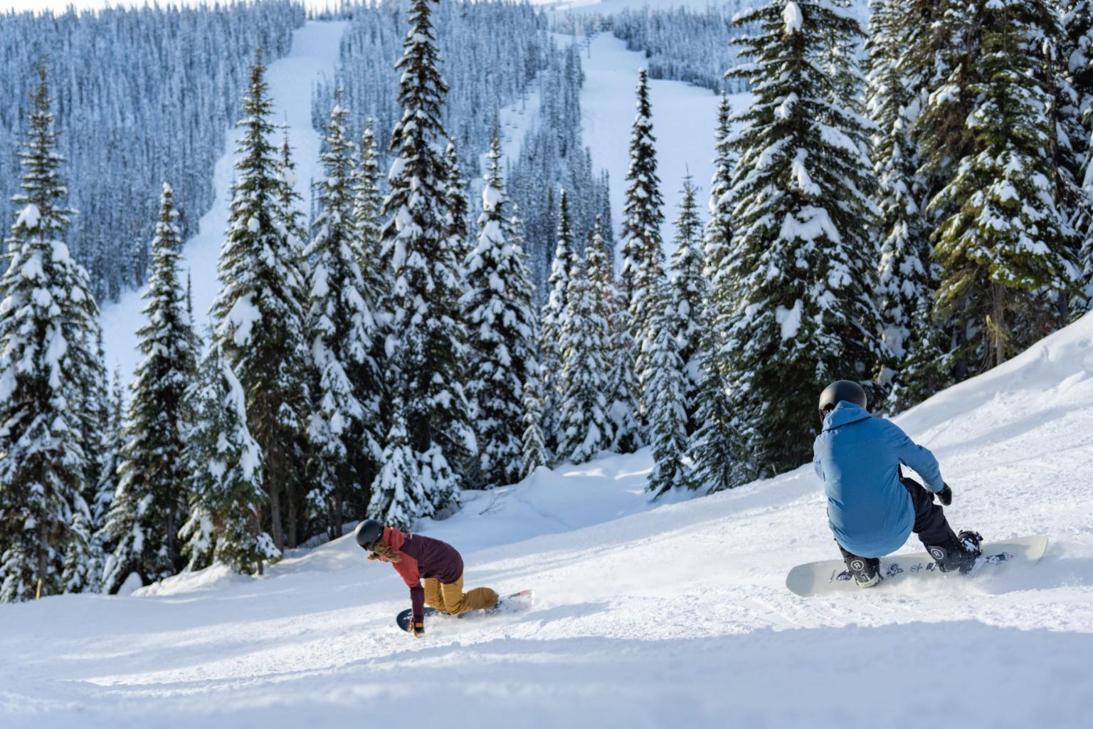Two people snowboarding down a run with mountains and trees in the background