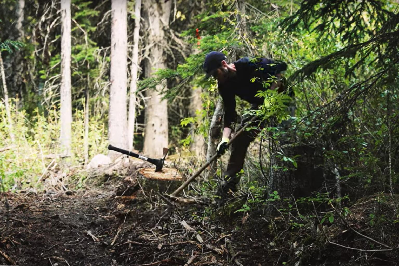 A trail builder digging on a trail with trees around him