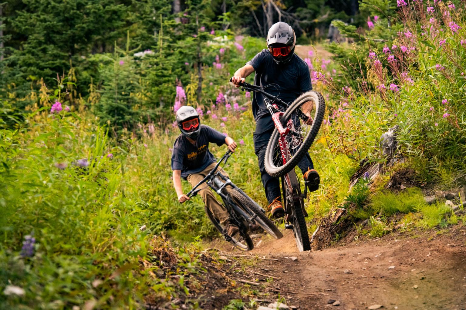 Two bikers hitting a bike jump on a trail surrounded by trees