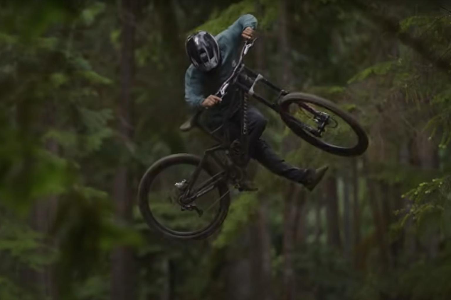 Biker hitting a jump on a DH bike with trees behind him